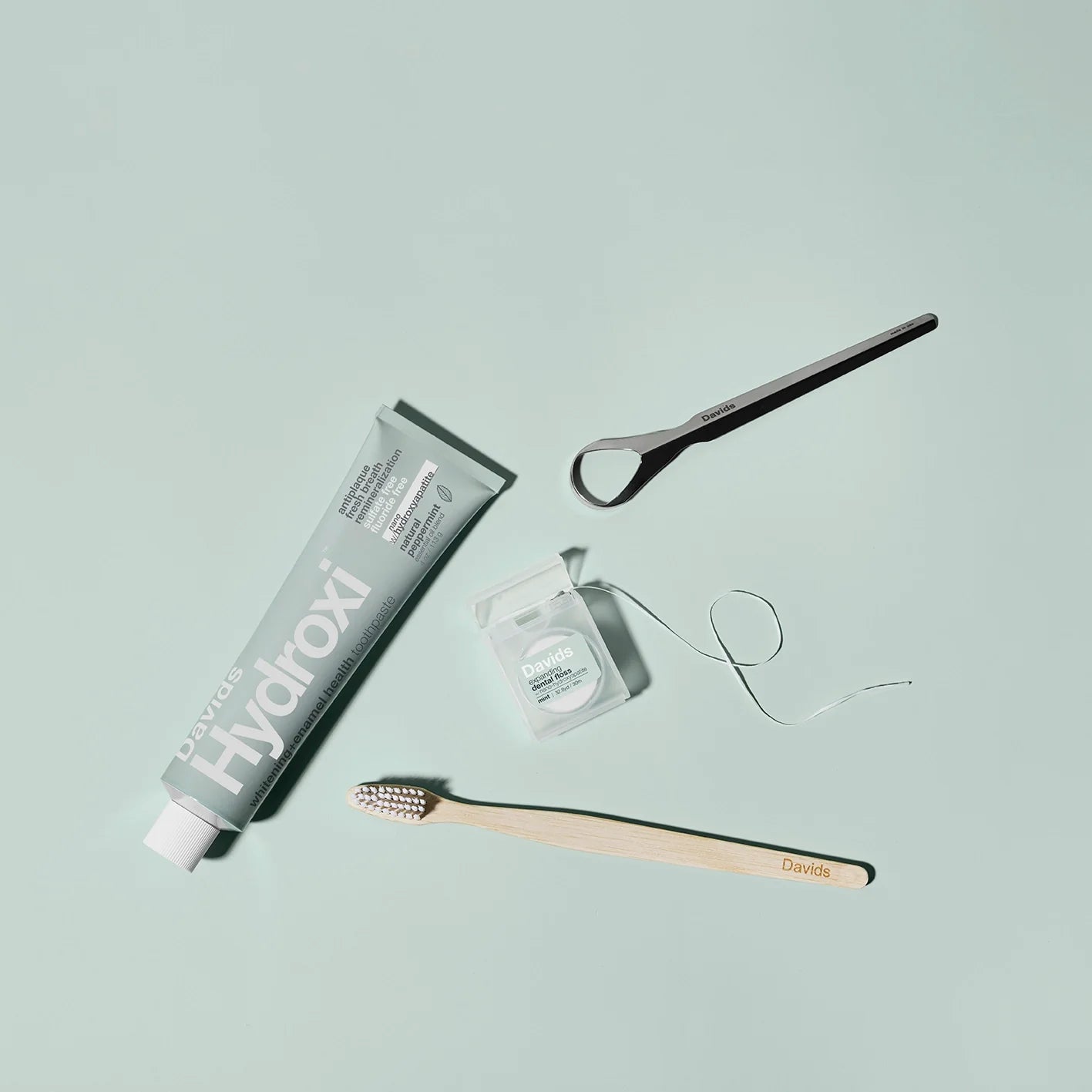 Davids Hydroxi™ balinamoji dantų pasta su nano hidroksiapatitu kartu su dantų siūlu, šepetėliu ir liežuvio valikliu, žaliame fone.