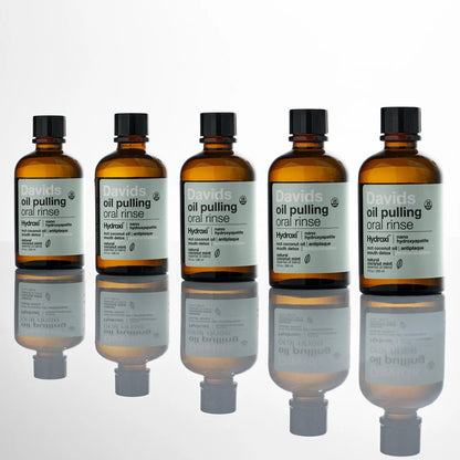 Davids Hydroxi™ burnos skalavimo skysčio aliejaus pagrindu buteliukai ant skaidraus paviršiaus su veidrodiniu atspindžiu.