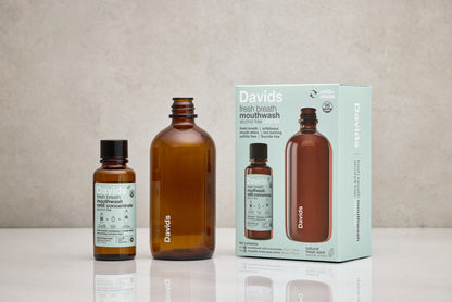 Davids Hydroxi natūralaus burnos skalavimo skysčio rinkinys ant stalo – daugkartinio naudojimo butelis, koncentratas ir pakuotė.